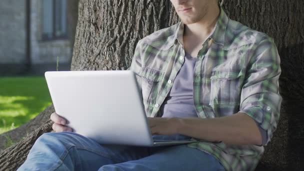 Fiatal férfi ült használ laptop, egyetemi hallgató dolgozik a projekt fa alatt — Stock videók