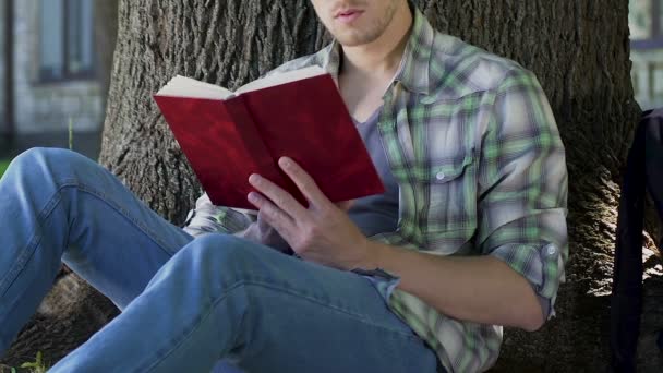 Guy leitura emocionante livro sob a árvore, absorvido em enredo, ávido leitor de livros — Vídeo de Stock