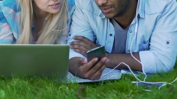 Guy dans les écouteurs avec téléphone, fille avec ordinateur portable couché ensemble sur l'herbe, amis — Video