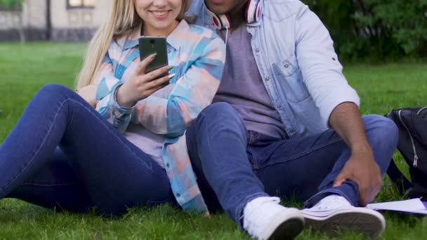 Couple sitting on lawn closely and looking at mobile phone of girl, gadgets — Stock Video