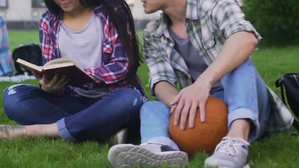 Studenten sitzen zusammen auf dem Rasen, Mädchen lesen und Mann spricht mit ihr, flirten — Stockvideo