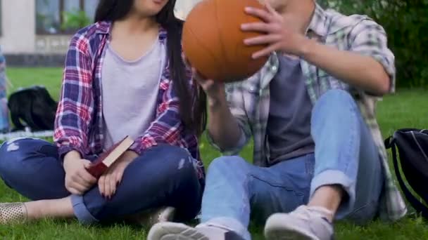 Девушка с книгой, парень с баскетбольным мячом, сидящий трава и говорить, отношения — стоковое видео