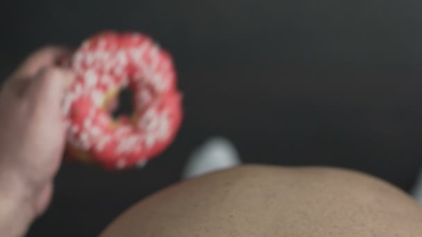 Вид сверху: мужчина с избыточным весом ест высококалорийный пончик с розовой каемочкой, ожирение — стоковое видео