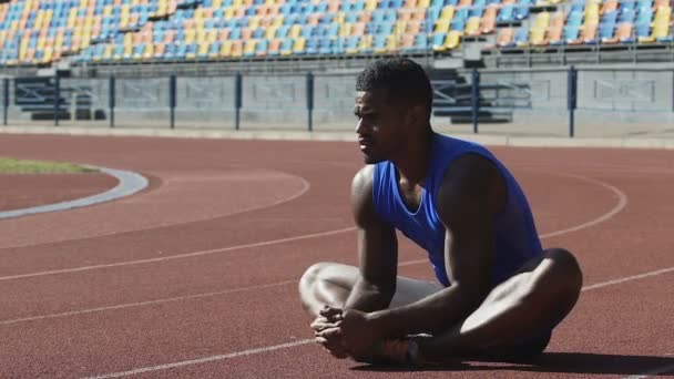 Fuerte atleta sentado en pista de atletismo haciendo ejercicios de calentamiento y flexibilidad — Vídeo de stock