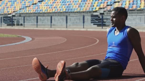 Homem bonito em roupas esportivas sentado na pista de corrida tomando fôlego e descansando — Vídeo de Stock