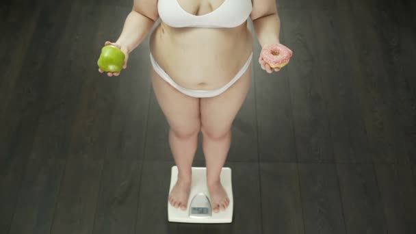 Избыточный вес женщины, стоящей на весах с яблоком и пончиком, выбор диеты — стоковое видео