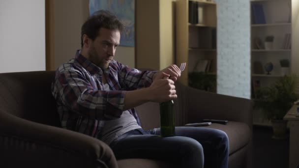 Man sitting on couch in dark room, trying to open beer bottle with fork, despair — Stock Video