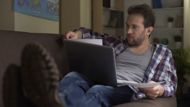 Man op de bank kijken naar papieren in handen met de laptop op schoot, betalen van rekeningen — Stockvideo