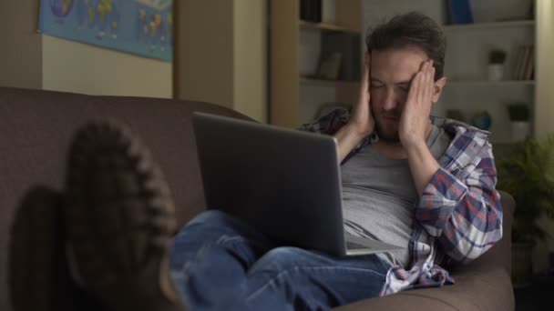 Cansado adulto macho no sofá olhando para laptop concentrado, busca de emprego contínua — Vídeo de Stock