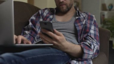Dizüstü bilgisayar ile kanepede oturan adam cezalandırmak veren cep telefonu ekran aşağı kaydırma