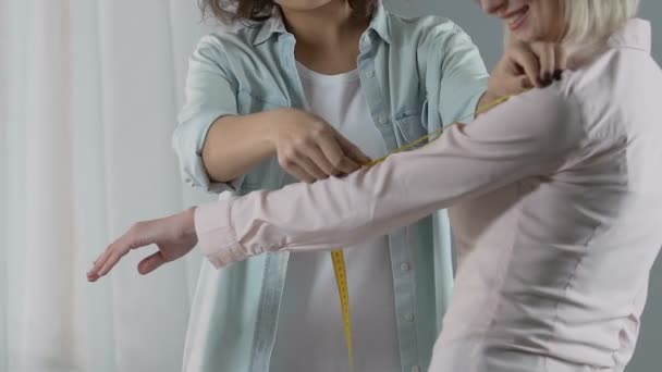 Улыбающийся дизайнер измеряет руку клиентки, обсуждая заказ одежды — стоковое видео