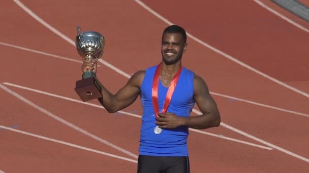 Muscular sportsman showing his medal and cup for victory to fans on stands — Stock Video