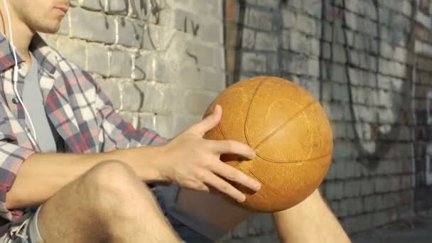 Jovem ouvindo música e girando bola cesta nas mãos, esperando amigos — Vídeo de Stock