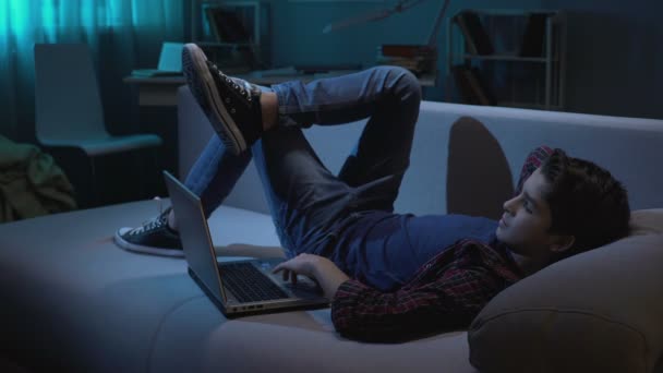 Чоловік студент розслабляється на дивані з ноутбуком, переглядаючи зображення в соціальній мережі — стокове відео