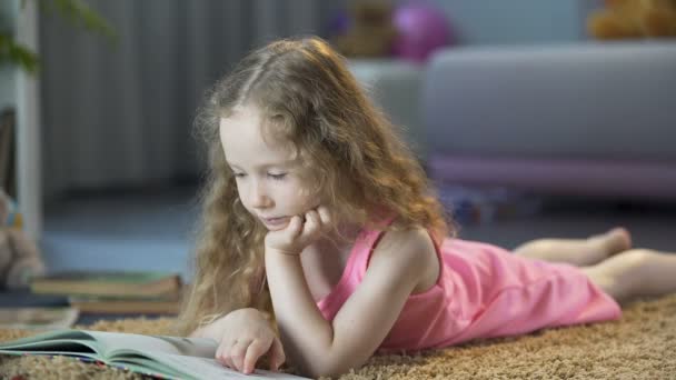 Niña curiosa tirada en el suelo, leyendo interesante libro de cuentos para niños — Vídeos de Stock