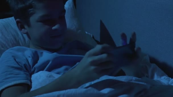 Возбужденный подросток играет в игру на смартфоне лежа в постели ночью, дисциплина — стоковое видео