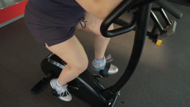 Chica regordeta motivada pedaleando rápido en bicicleta estática en el gimnasio, entrenamiento activo — Vídeos de Stock