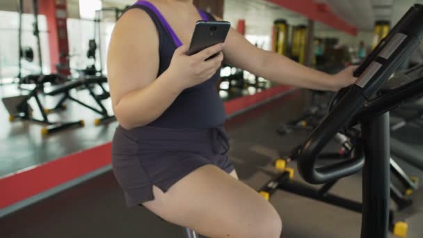 Llenita hembra montando perezosamente bicicleta de ejercicio y viendo vídeo en el teléfono celular — Vídeos de Stock