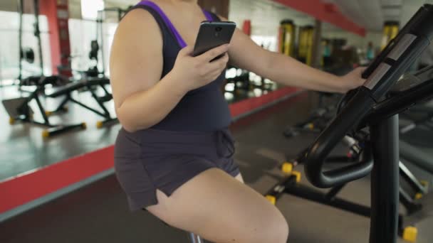 Plump female riding sluggishly exercise bike and watching video on cellphone — Stock Video