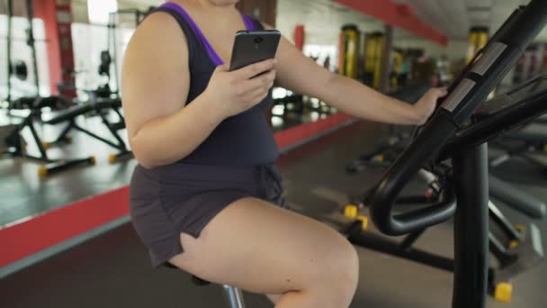 Mujer con sobrepeso montando lentamente bicicleta estacionaria y charlando en el teléfono móvil — Vídeos de Stock