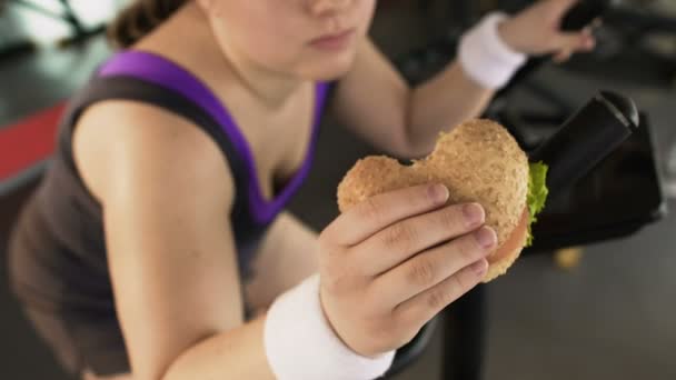 Товста жінка катається на стаціонарному велосипеді та їсть смачний бургер у тренажерному залі, здорове харчування — стокове відео