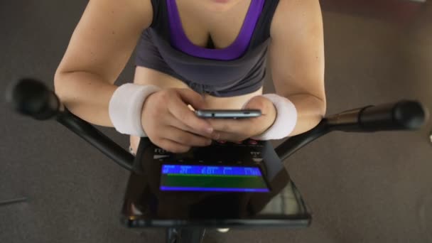 Mujer obesa desplazándose en el teléfono celular mientras monta bicicleta estática, aplicación de fitness — Vídeos de Stock