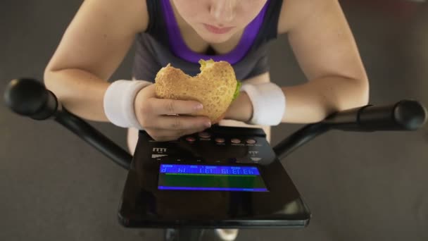 Жінка їсть жирну бургер під час їзди на стаціонарному велосипеді, фаст-фуді — стокове відео