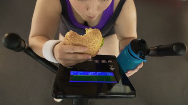 Knubbig flicka överätande burger och dricksvatten medan ridning motionscykel — Stockvideo