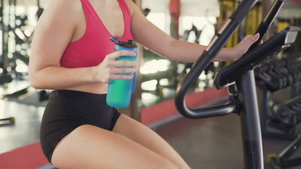 Entrenamiento en el gimnasio, mujer delgada beber agua mientras monta bicicleta estática, fitness — Vídeos de Stock