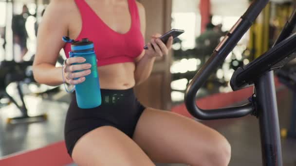 Young healthy female riding exercise bike and scrolling fitness app on cellphone — Stock Video