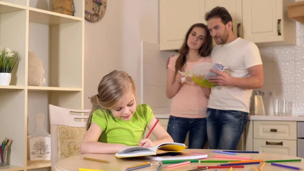 Pais felizes orgulhosamente assistindo sua filha inteligente fazendo lição de casa, família — Fotografia de Stock