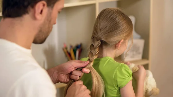 Ответственный хороший отец плетет плетеные волосы маленьких дочерей, готовится к школе — стоковое фото