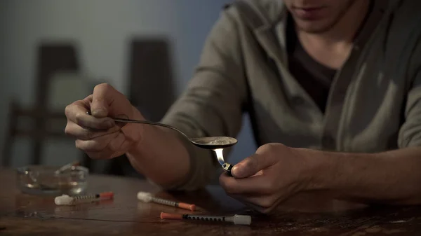 Maschio tossicodipendente che prepara la dose di eroina in cucchiaio con accendino, siringhe sul tavolo — Foto Stock