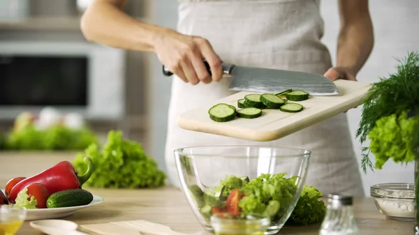 Cocinera profesional agregando rodajas de pepino fresco en un tazón de vidrio con ensalada —  Fotos de Stock