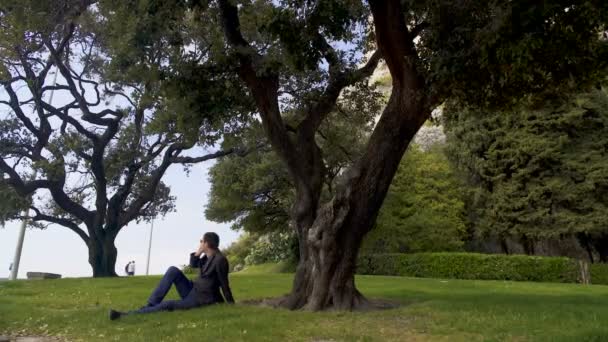 Занятой человек в классическом костюме расслабляется возле огромного дерева и разговаривает по телефону, панорама — стоковое видео