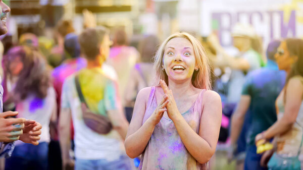 Excited young woman covered in Holi colors, dancing in crowd at concert, fun