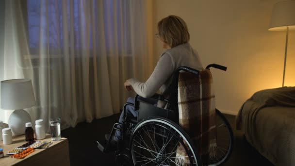 Инвалидная женщина в инвалидной коляске смотрит в окно на улицу, одиночество — стоковое видео