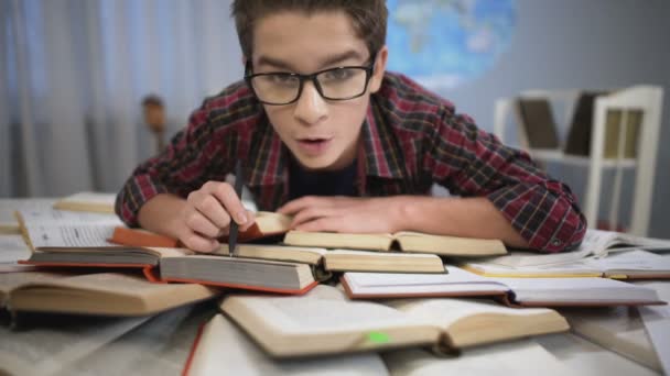 Niño estudiando cuidadosa y atentamente la literatura, gran pila de libros sobre la mesa — Vídeos de Stock