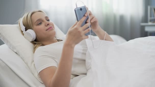 Adolescente en auriculares escuchando música en la cama, comunicándose en las redes sociales — Vídeos de Stock