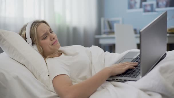 Upset teenage girl in headphones communicating in social networks, lying in bed — Stock Video