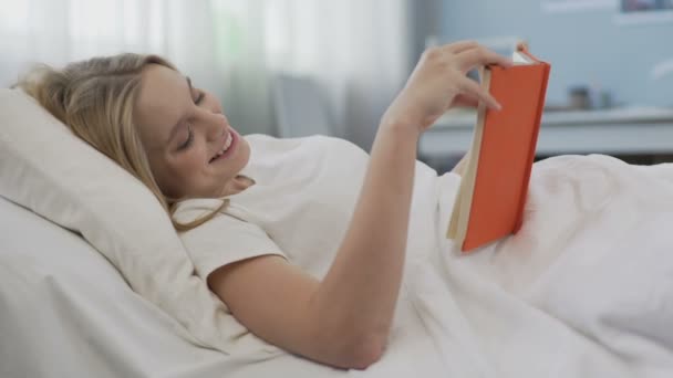 Smiling delighted teenage girl reading novel about romantic love, lying in bed — Stock Video
