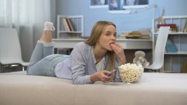 Tienermeisje kijken tv in de kamer met afstandsbediening in de hand en het eten van pop corn — Stockvideo