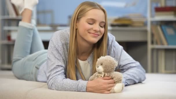 Улыбающаяся школьница обнимает свою игрушку плюшевого мишку в спальне, домашний уют — стоковое видео