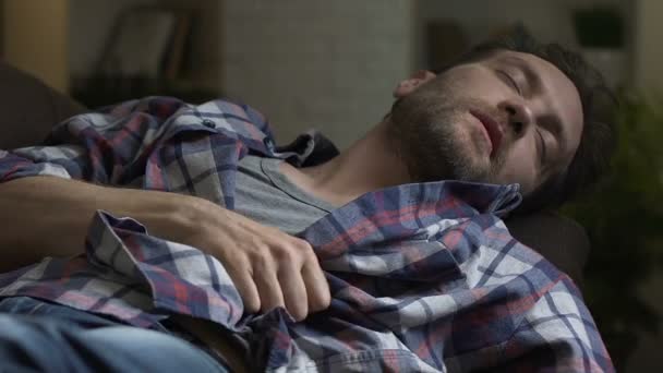 Неопрятный алкоголик спит на диване после домашней вечеринки, пьяный мужчина чешет живот — стоковое видео