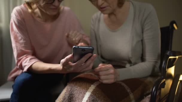 Είδος θηλυκό εθελοντών δείχνει ηλικιωμένη γυναίκα σε αναπηρική καρέκλα, πώς να χρησιμοποιείτε το smartphone — Αρχείο Βίντεο