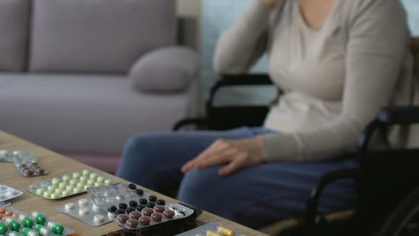 Alte kranke Frau im Rollstuhl schaut auf Tabletten auf dem Tisch und seufzt, Einsamkeit — Stockvideo