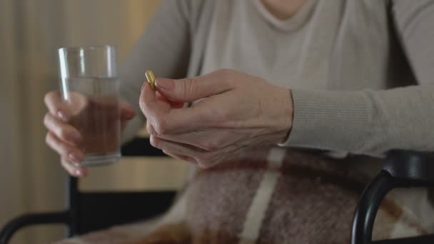 Mujer madura tomando píldoras para el reumatismo sentado en silla de ruedas, debilidad en las piernas — Vídeo de stock