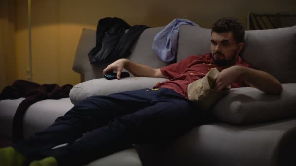Ленивый неряшливый холостяк смотрит телевизор, лежащий на диване после работы и ест чипсы — стоковое видео