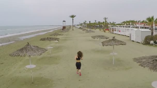 Attiva atletica femminile che corre sulla spiaggia tra ombrelloni di paglia, vista aerea — Video Stock