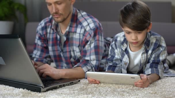 Pequeno filho jogando no tablet no chão em casa, enquanto o pai trabalhando no laptop — Vídeo de Stock
